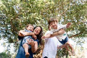 Parents playfully holding and swinging two small children outdoors amongst green trees on a sunny day.