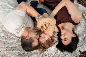 Three people lying on a blanket together holding hands and laughing while looking up at the sky.