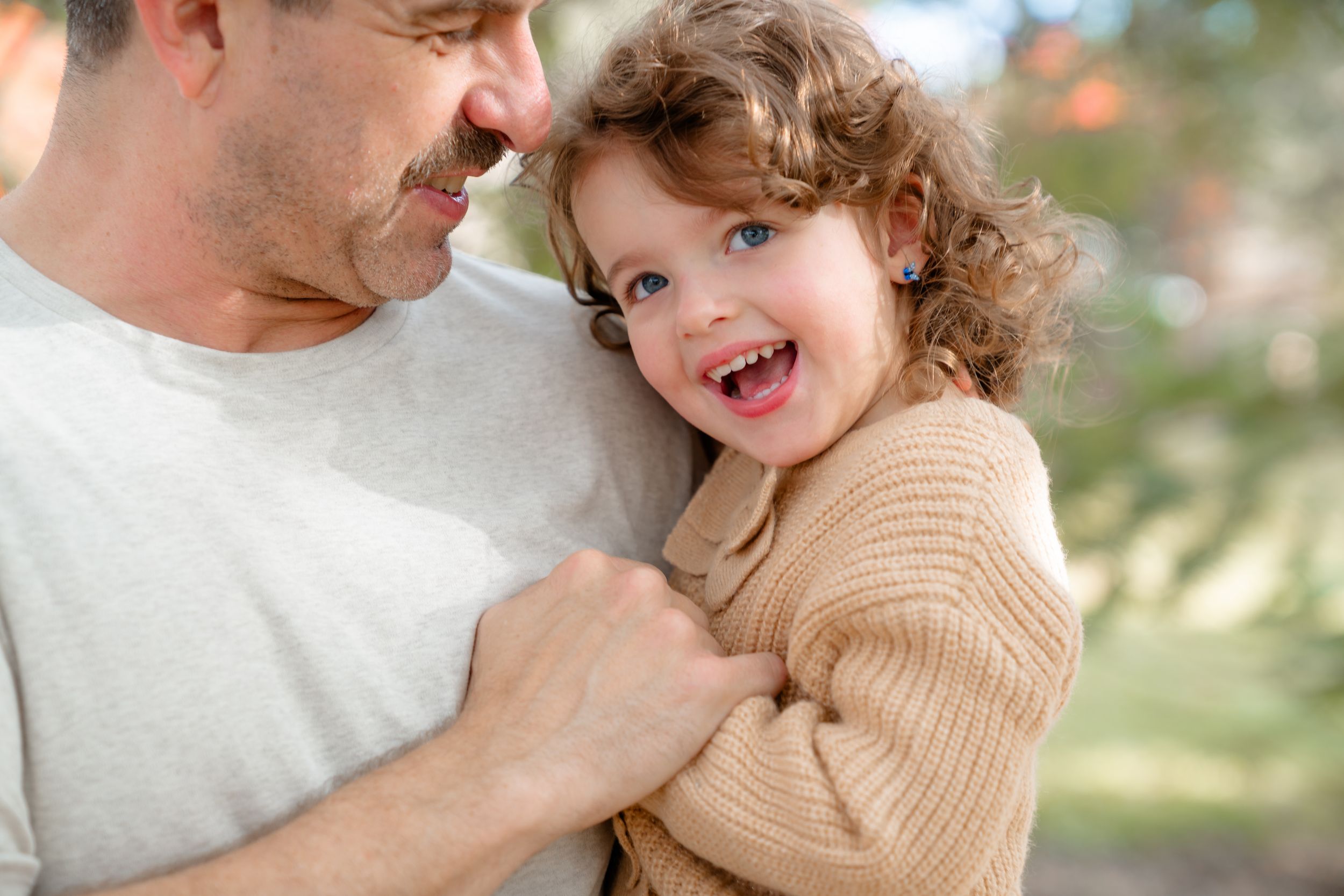 A happy child with curly hair wearing a tan sweater shares a joyful moment with someone in a white shirt outdoors.