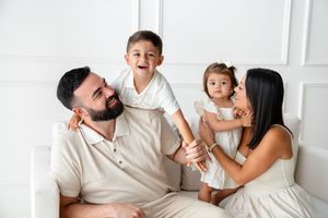 A family dressed in white sits together indoors, sharing joyful laughter and embraces against a light backdrop.