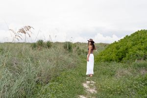 A person in a white dress walks through tall grass along a coastal path on a sunny day.