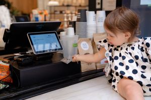 A small child in a polka dot dress reaches over a retail counter towards a touchscreen point-of-sale system.