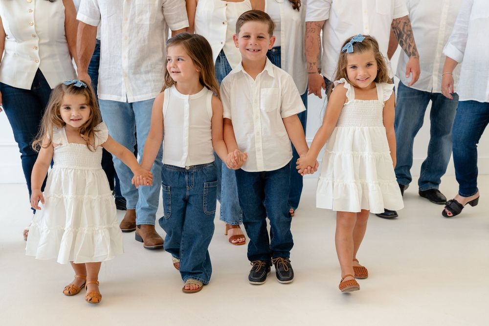 Four young children in white outfits and jeans hold hands walking together during a family photo session.