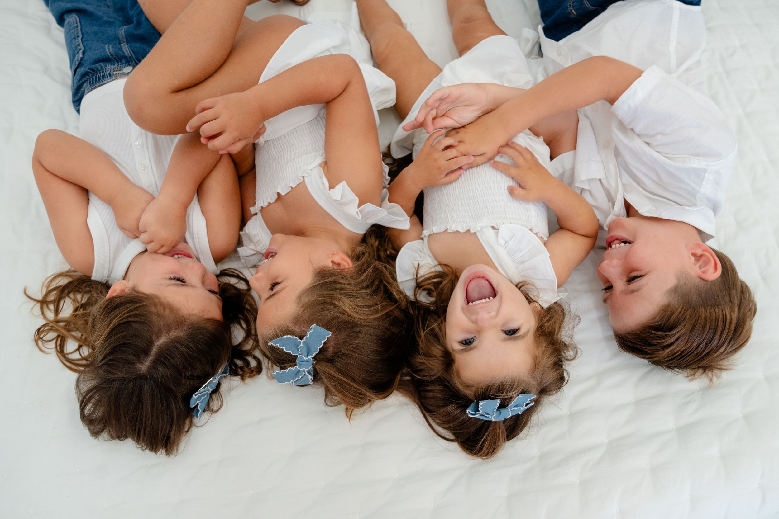 Four happy children lying on a white bed laughing together, wearing white outfits with blue hair bows.