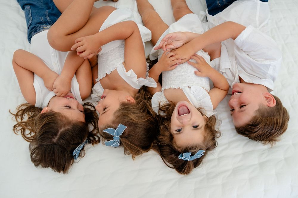 Four happy children lying on a white bed laughing together, wearing white outfits with blue hair bows.