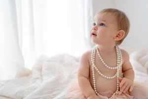 A baby wearing pearl necklaces sits in a tutu on white bedding while looking up with wonder.