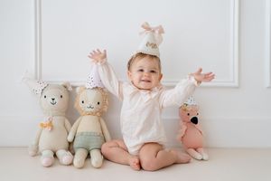 Baby in white outfit sits on floor surrounded by soft plush animal toys in pastel colors against white wall.