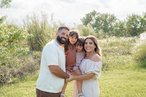 A family stands together outdoors in a sunny field with tall grass and trees in the background.