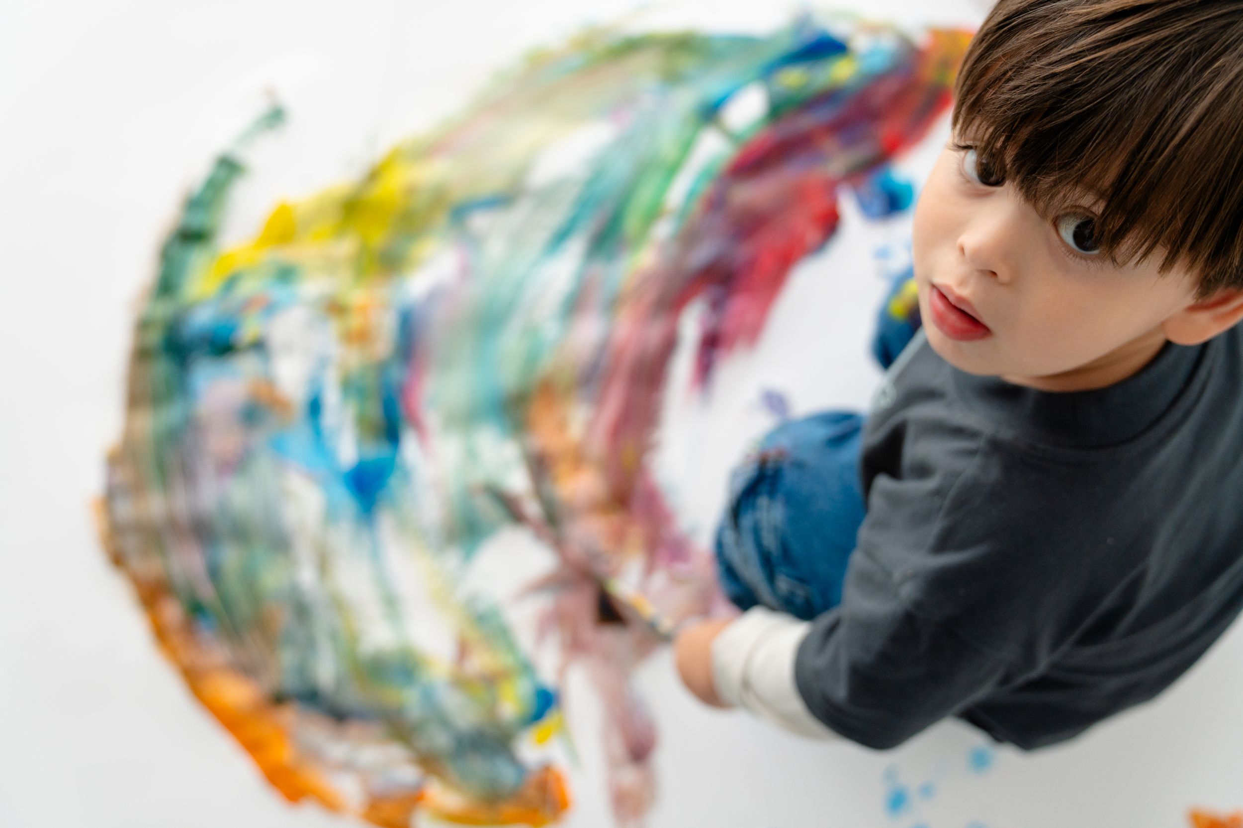 Young boy painting colorful abstract art on white canvas, viewed from above, wearing dark blue shirt.