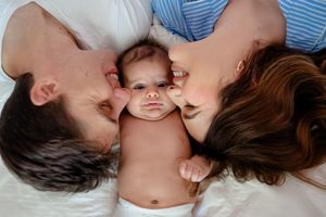 Parents kissing their newborn baby on the cheeks while lying on a white bed together.