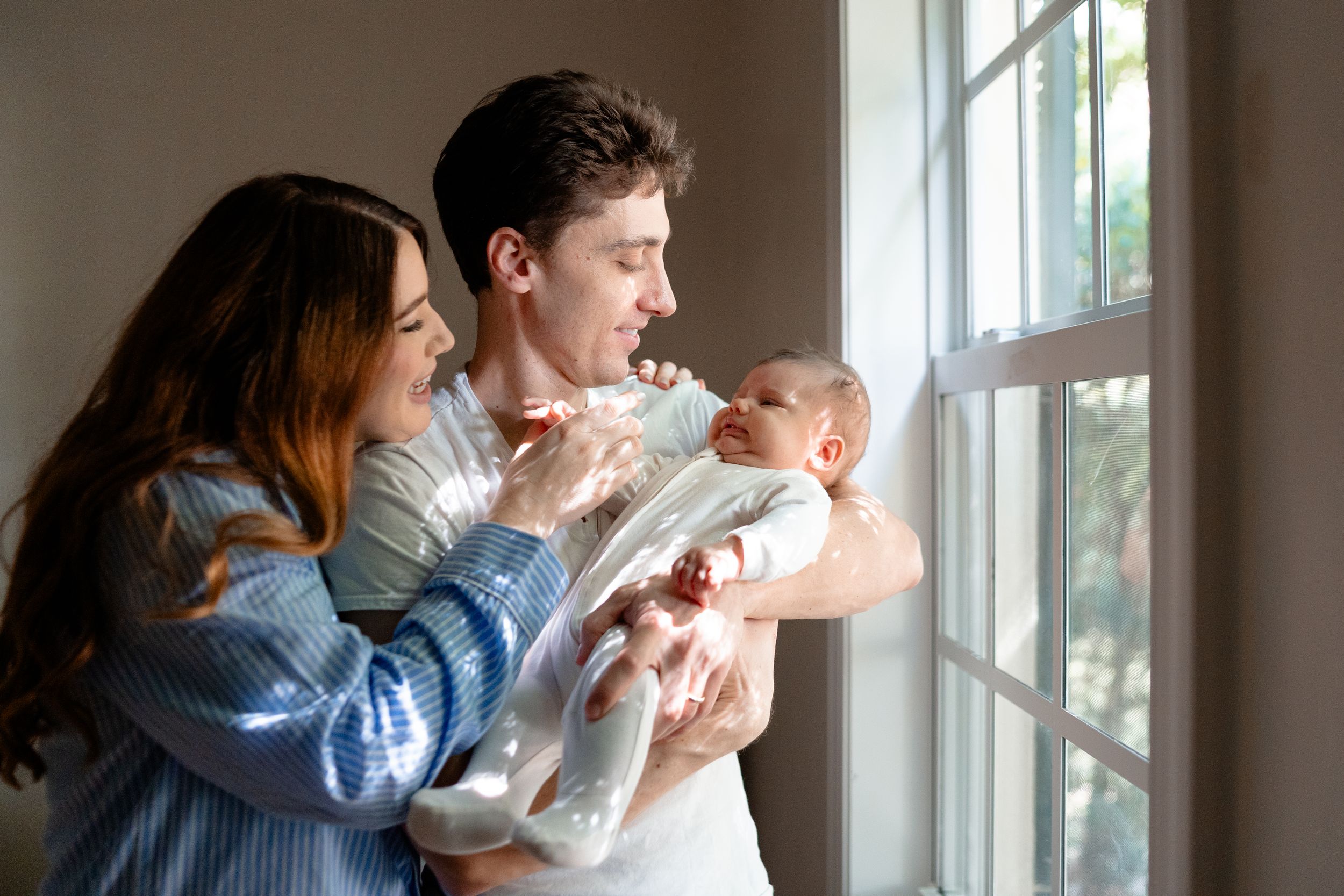 Young couple smiling and holding newborn baby near a bright window in a cozy home setting.