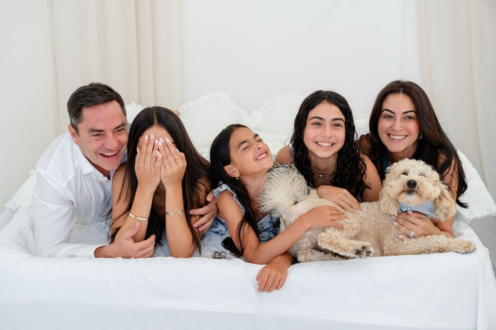 Happy family of four with a fluffy dog lying on a white bed, smiling and laughing together indoors.