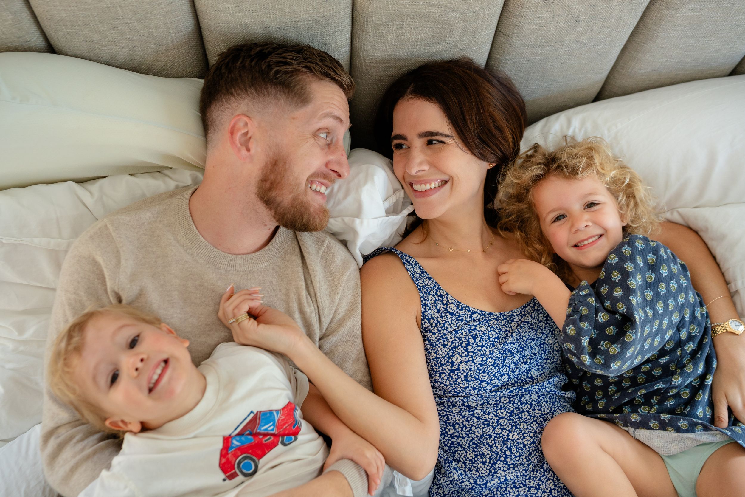 Happy family of four lying on a bed, parents smiling at each other with two young curly-haired children.