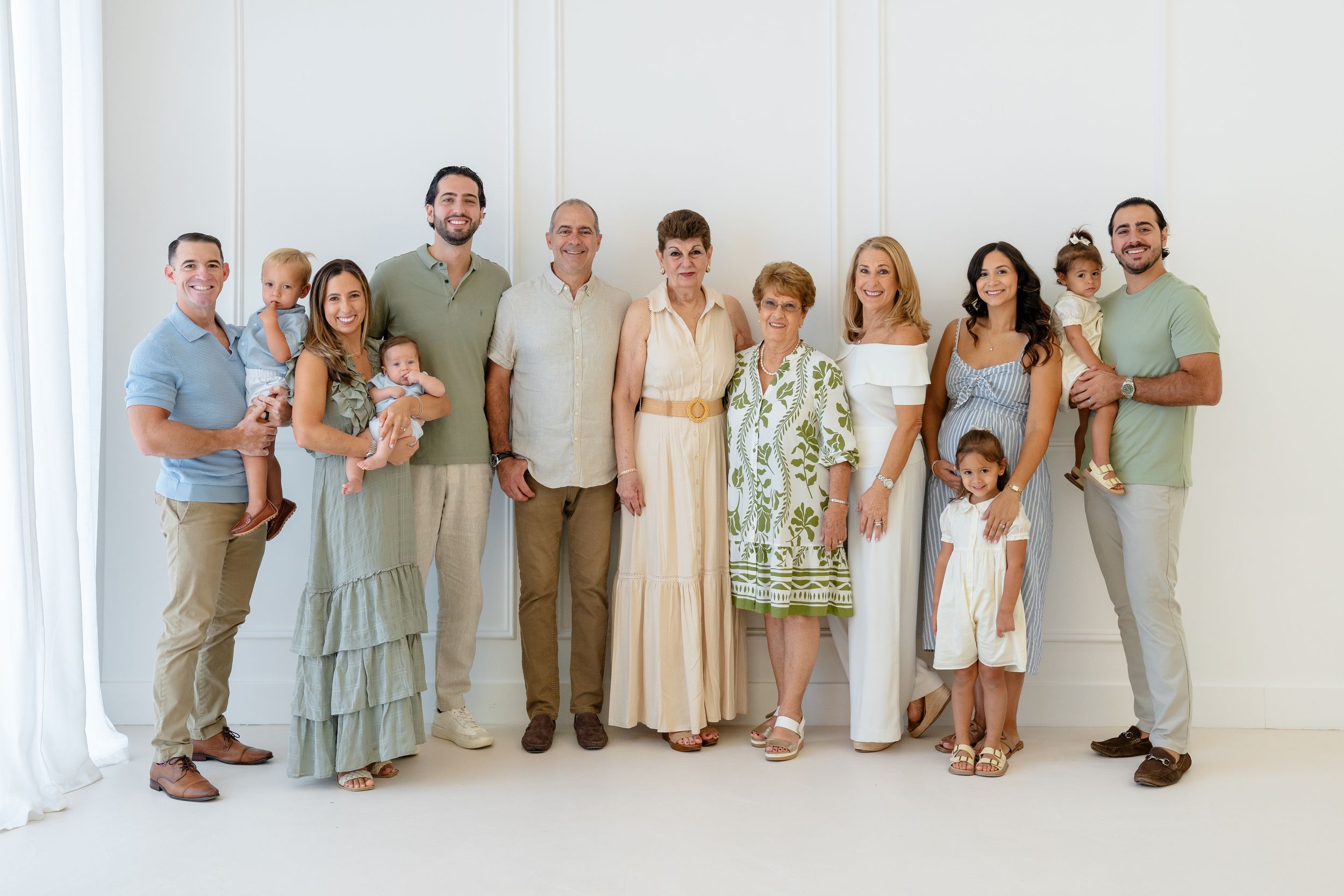 Large multigenerational family portrait posing together in a bright white studio, dressed in neutral and pastel tones.