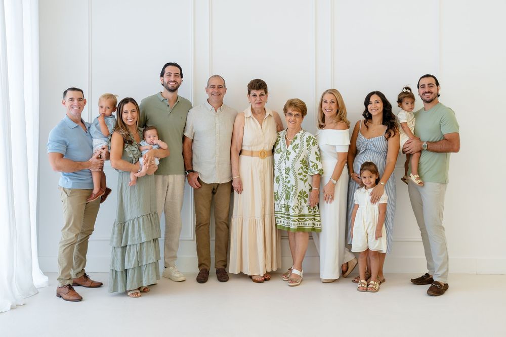 Large multigenerational family portrait posing together in a bright white studio, dressed in neutral and pastel tones.