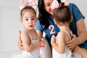 Two toddlers in white dresses and pink bows join a parent seated on the floor showing birthday number two decoration.