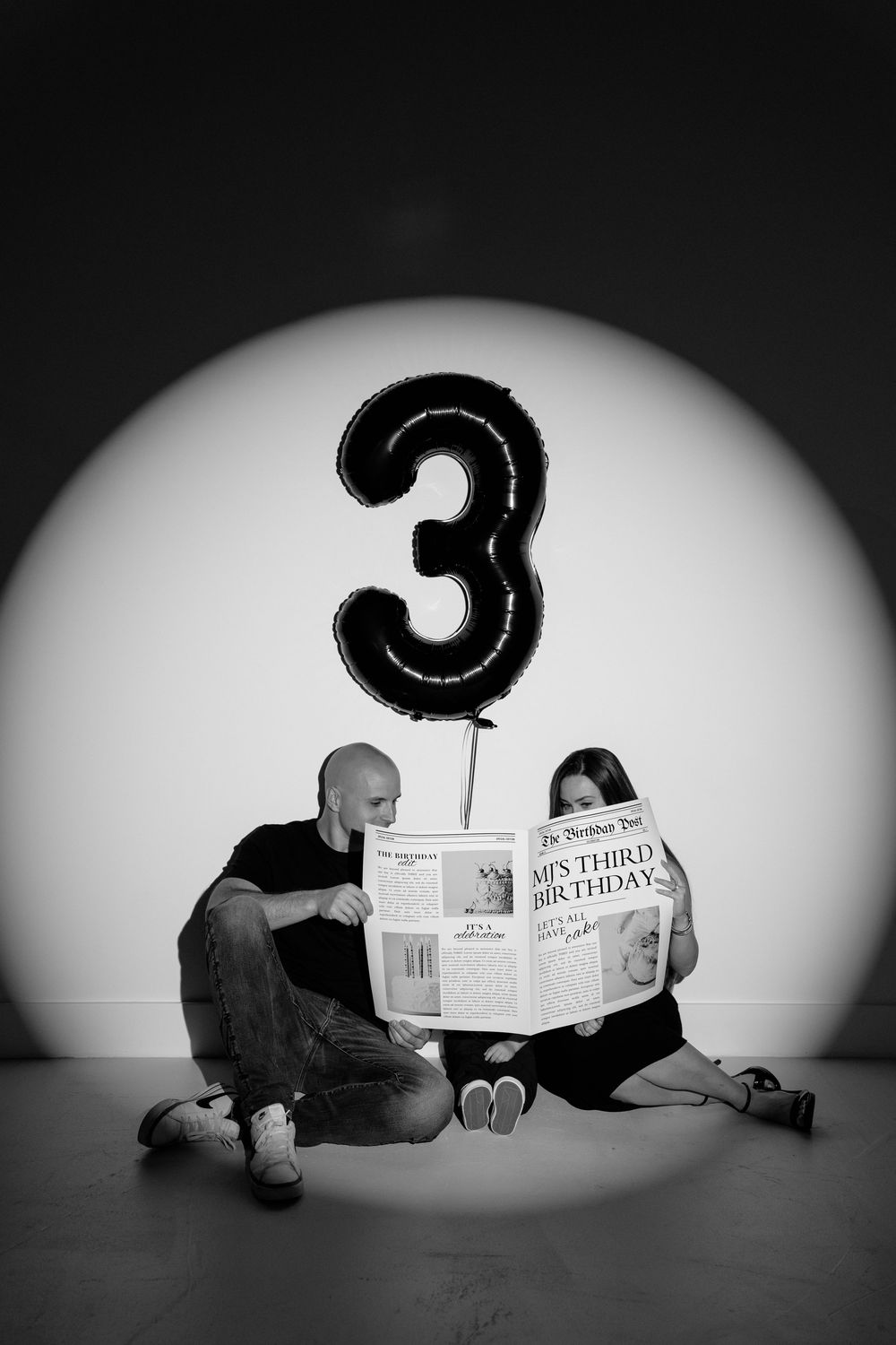 Black and white photo of couple sitting on floor reading newspaper beside number 3 balloon celebrating a third birthday.