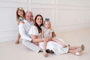 A parent and child sit casually together on a white floor in coordinated white outfits.