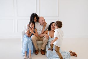 A joyful family shares a candid laughing moment together while sitting on the floor in a bright white room.