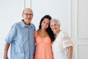 Three generations stand together smiling against a white wall, with coral and light blue clothing.