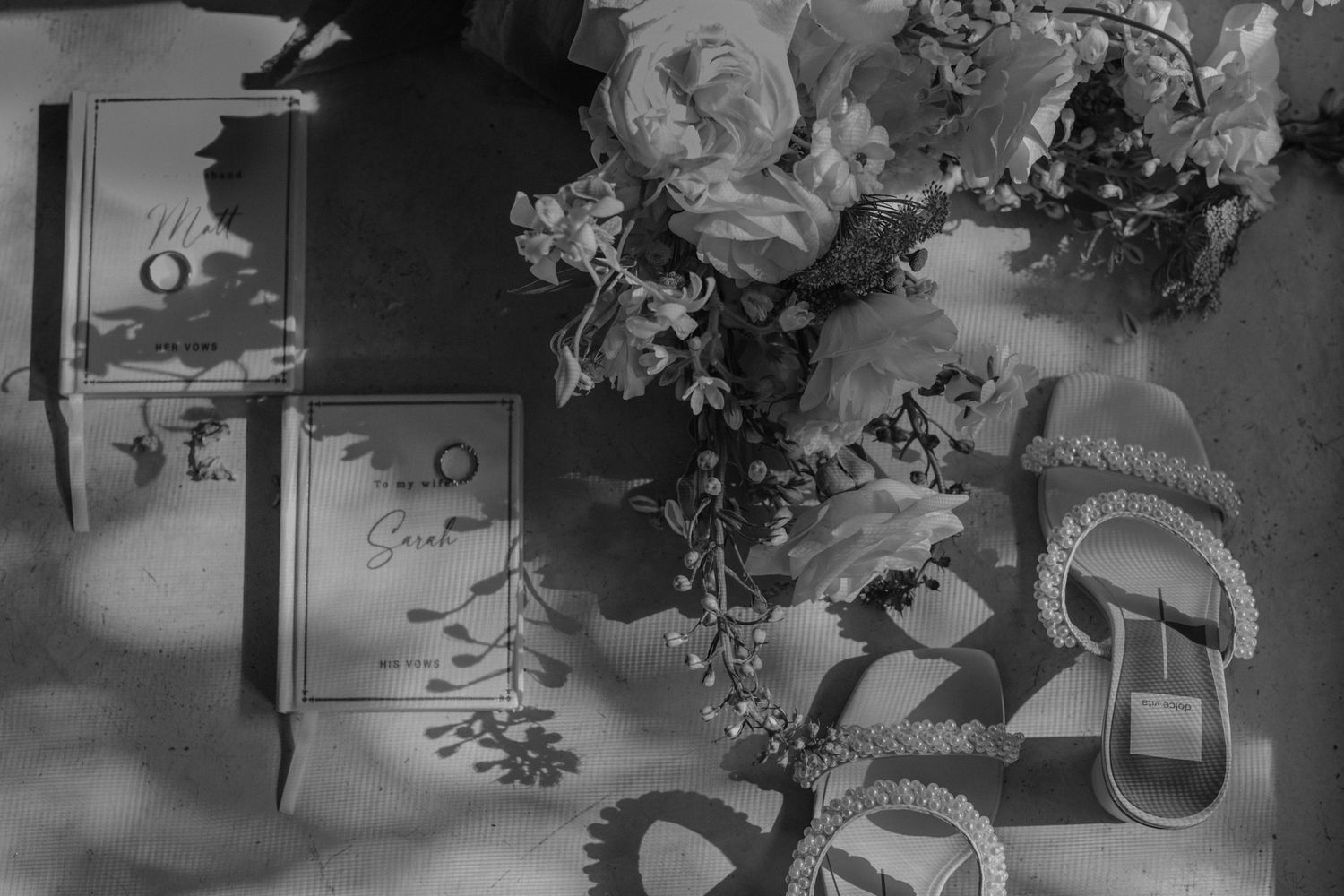 Black and white flat lay of wedding stationery, floral bouquet, and embellished sandals on a textured surface.