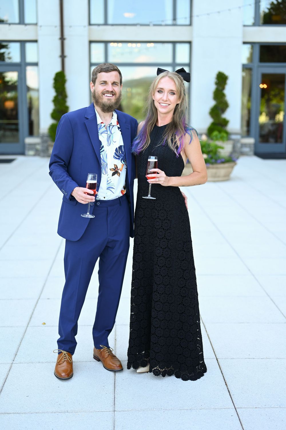 A couple in formal attire poses with wine glasses on a modern white patio.