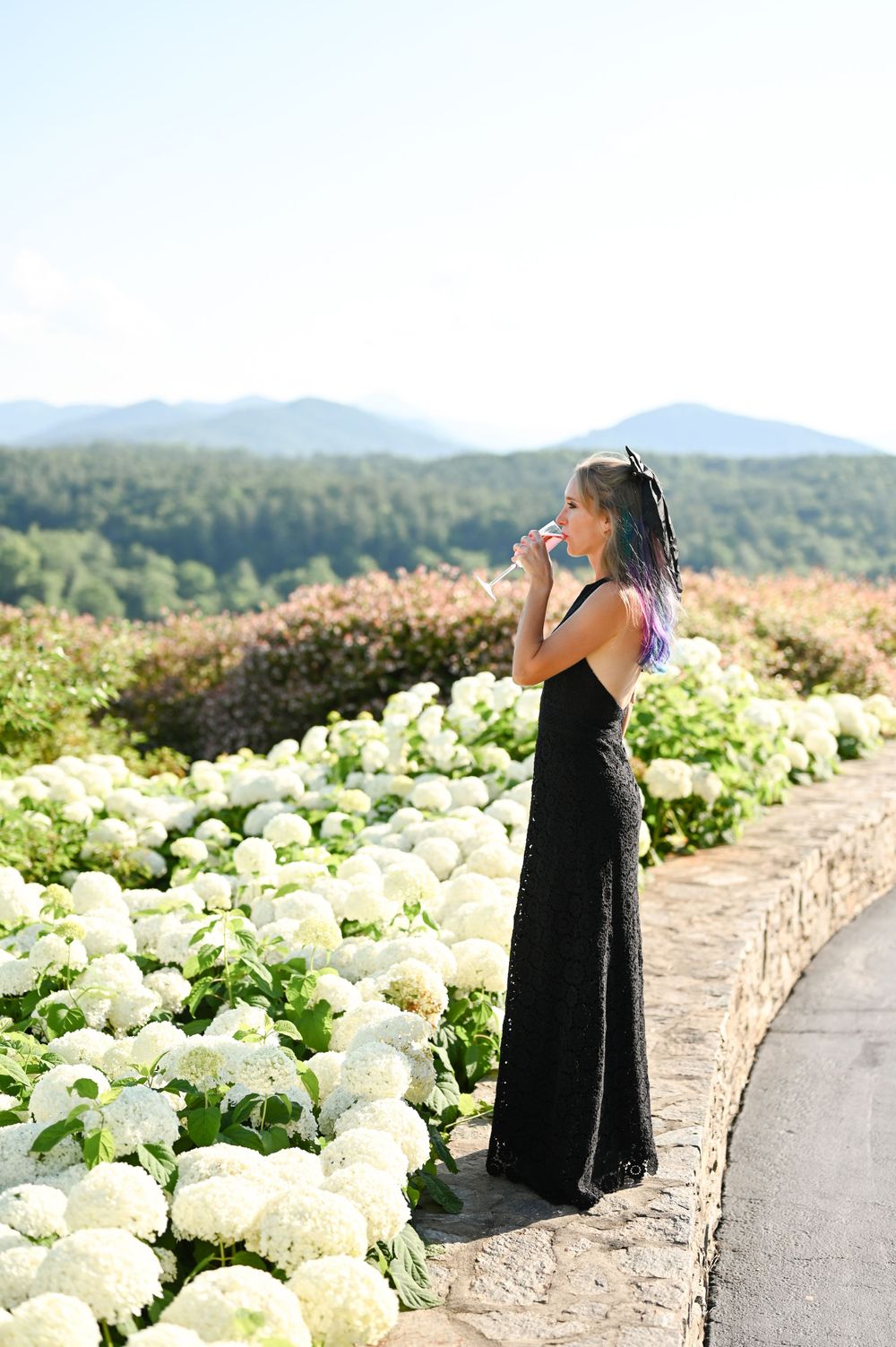 A figure in a black dress poses along a garden path lined with white hydrangea flowers and mountain views.