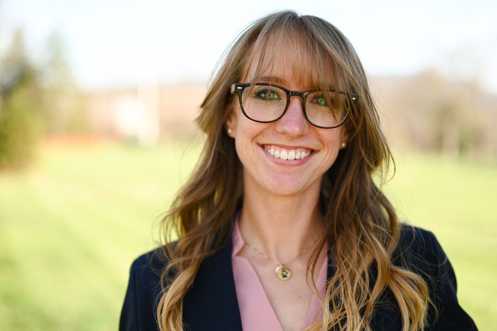 Professional headshots of someone wearing glasses and a blazer against a blurred natural background.