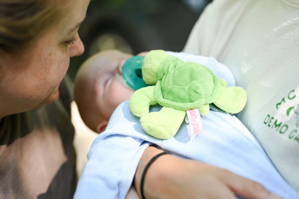 Baby in blue outfit holding a small green plush toy while being cradled.