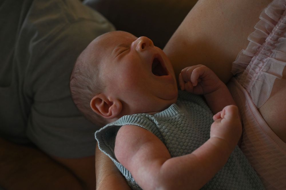 A baby in a green knit onesie yawns while lying down during a peaceful moment.