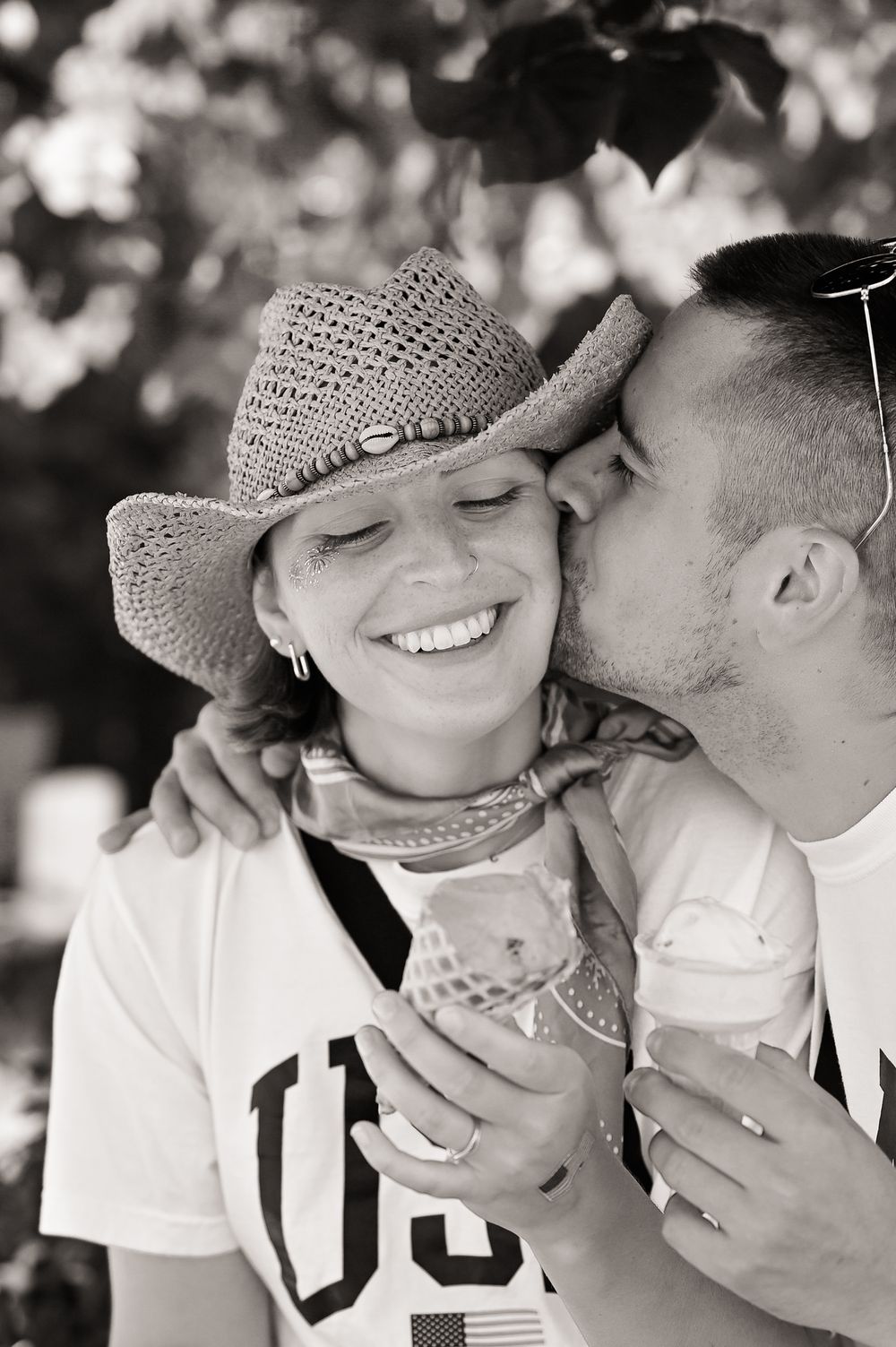 Black and white candid photograph showing a couple sharing a tender moment outdoors, one wearing a straw cowboy hat.