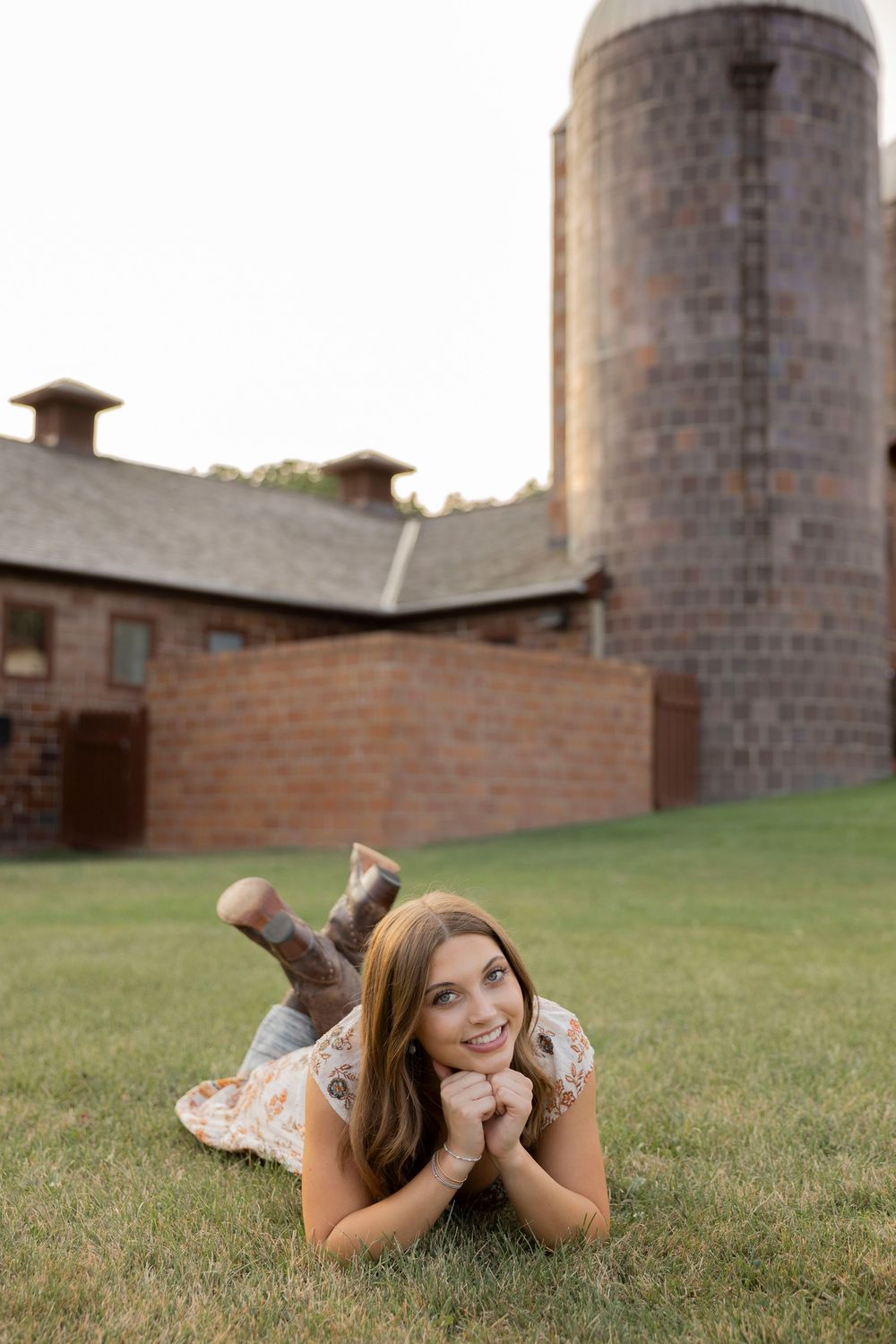 senior in high school in floral dress lying on grass, smiling, with rustic brick barn and silo in background at van hoosen farms