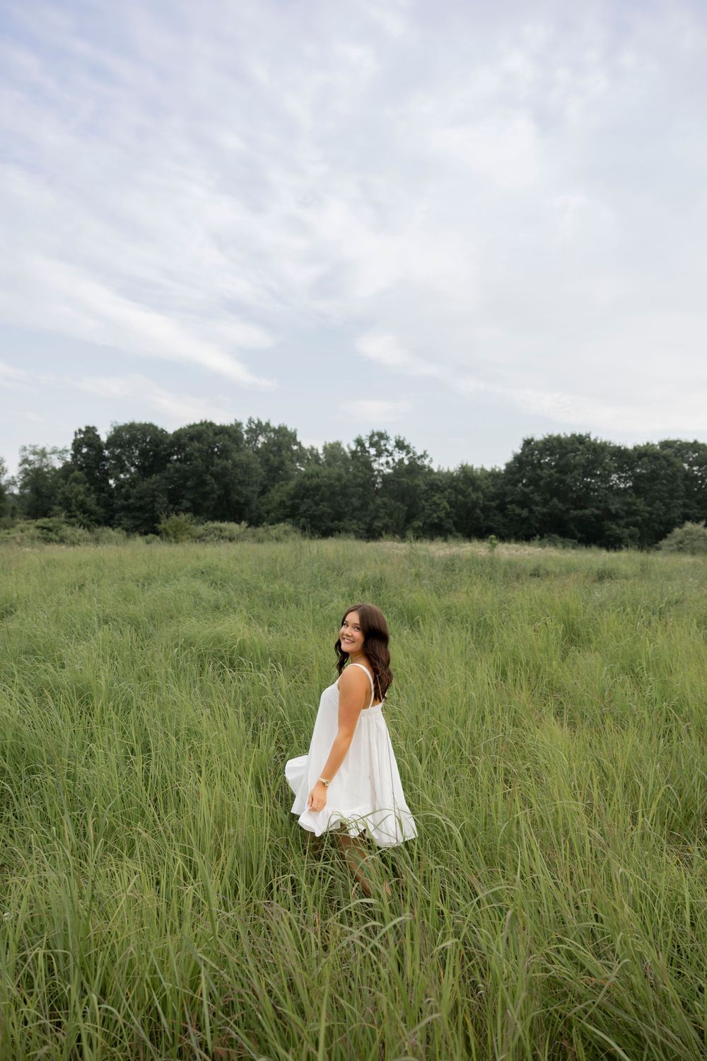 A young woman in a white sundress walks through tall green grass in a lush meadow with trees in the background at stoney creek metropark