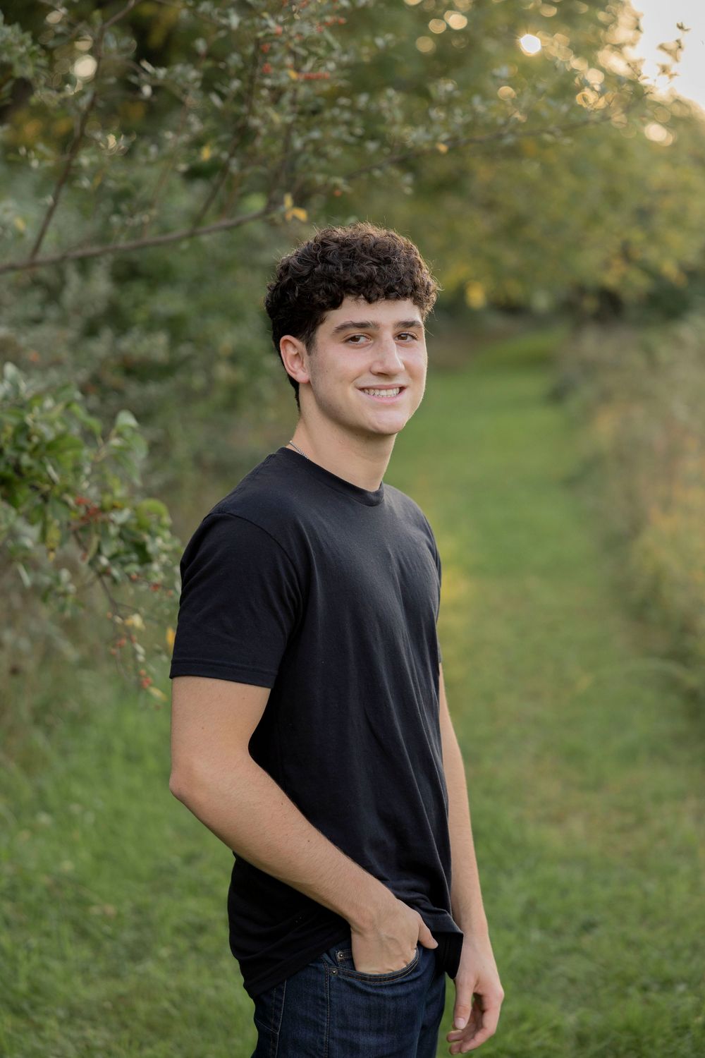 Young man in black t-shirt and jeans smiling outdoors on a green nature path during golden hour at troy farm park in michigan