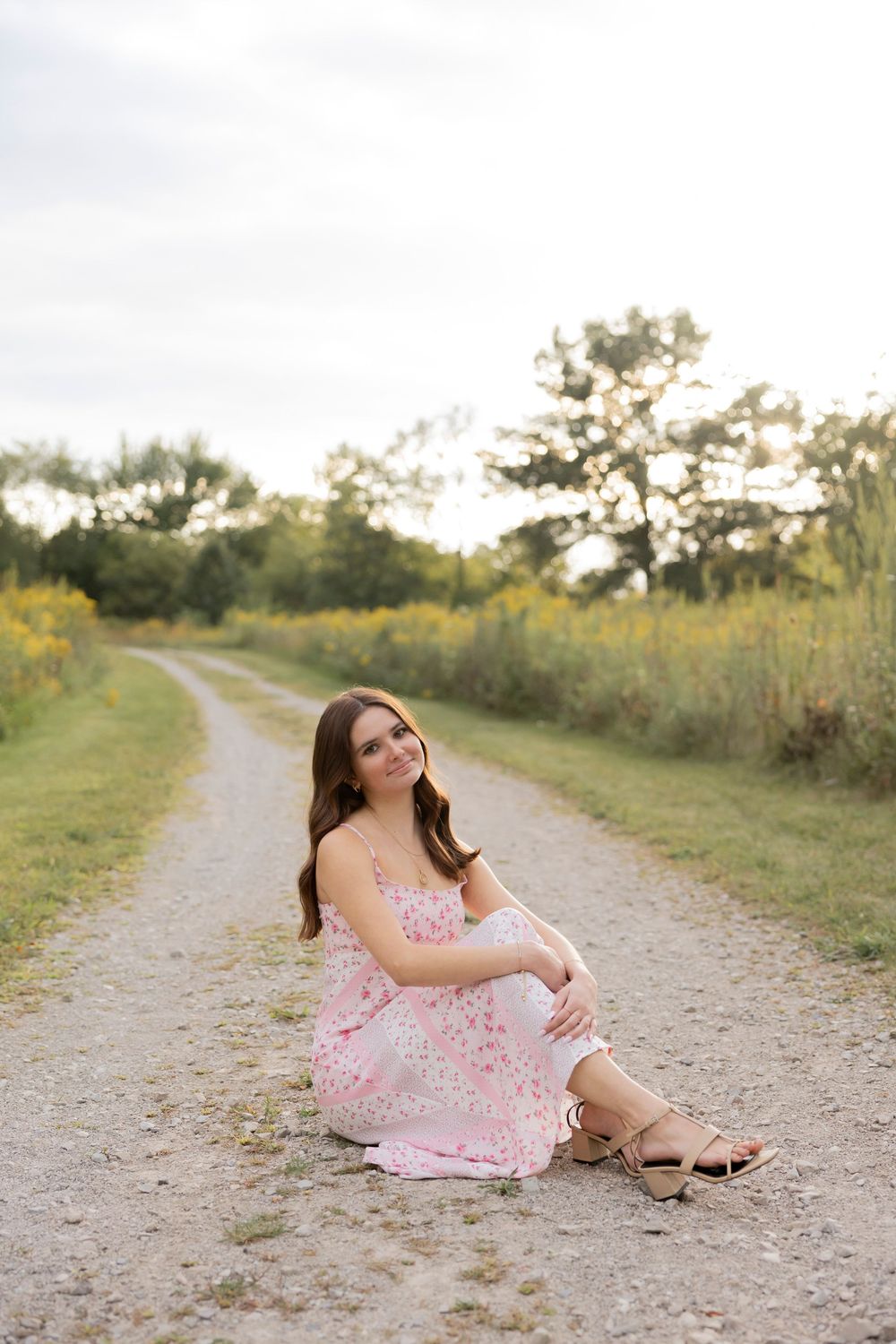 high school senior in a pink floral dress poses gracefully on a winding gravel path surrounded by wild greenery in Rochester hills michigan