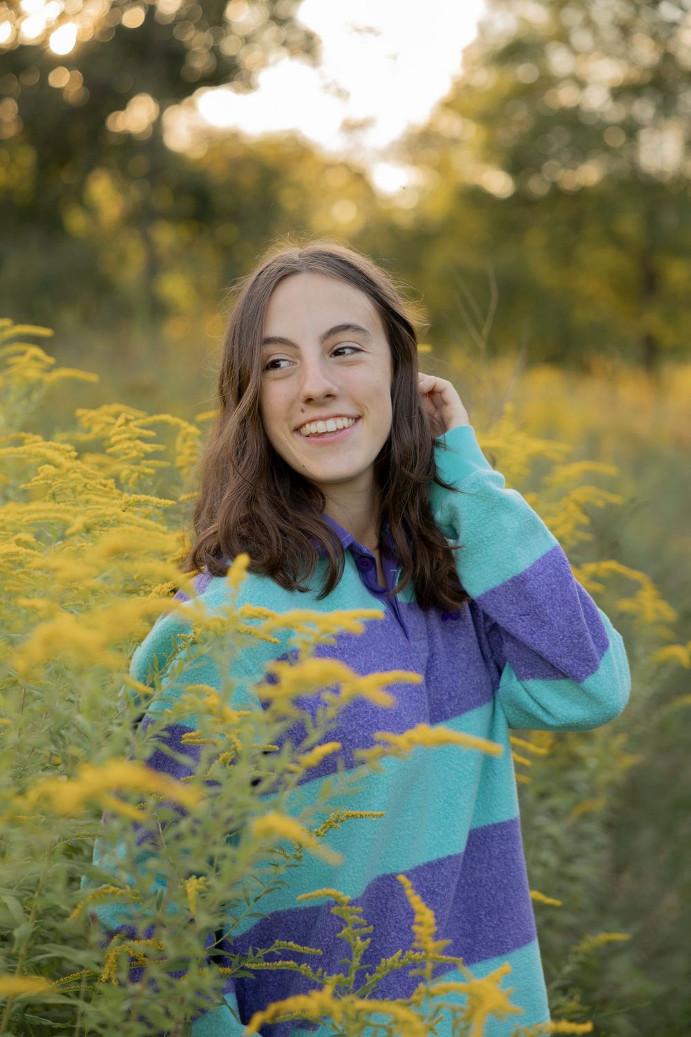 A high school senior wearing a vibrant purple and turquoise sweater poses playfully in a field of yellow wildflowers at Addison oaks park