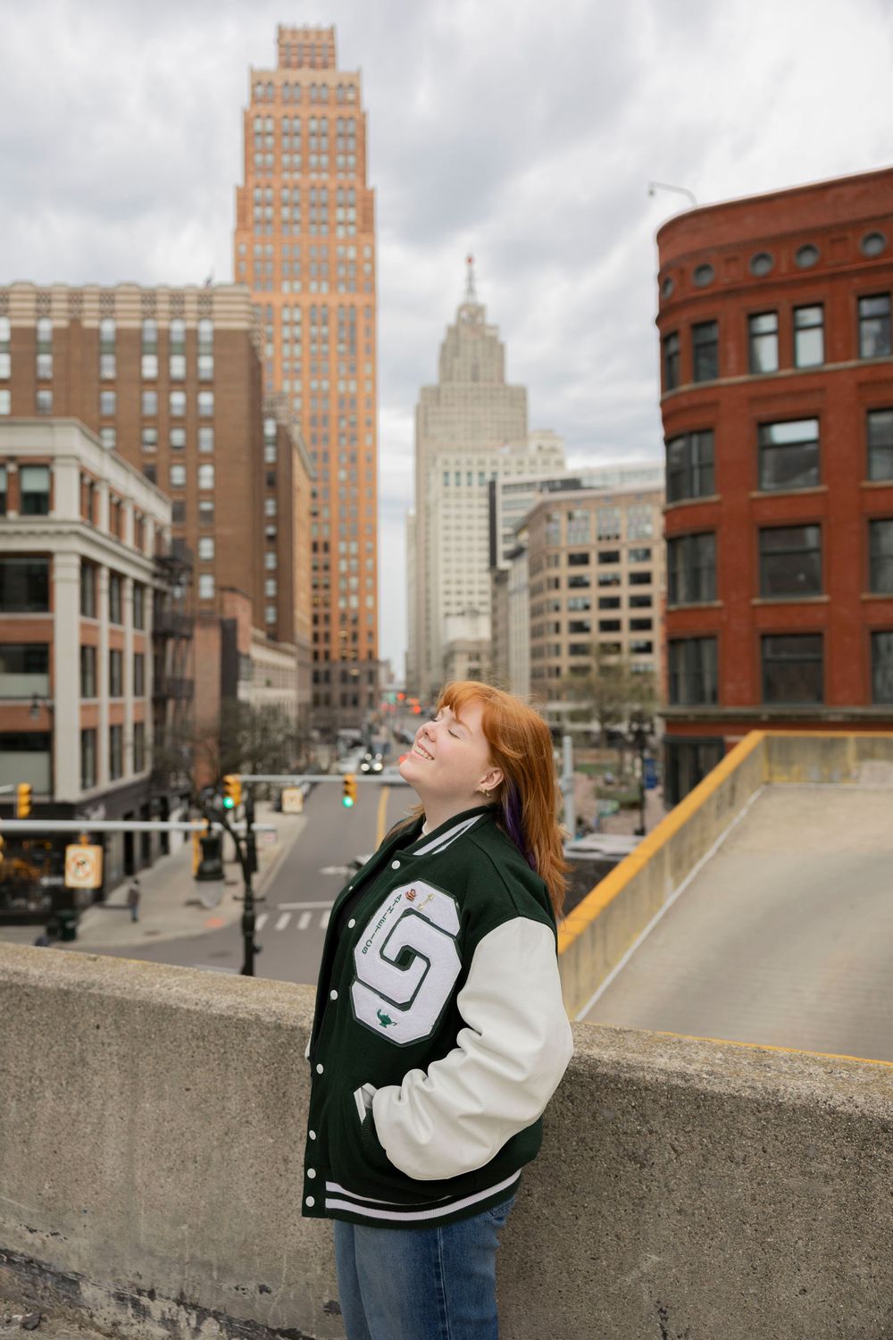 high school senior in lake Orion varsity jacket poses on urban rooftop with Detroit skyline and tall buildings in background.