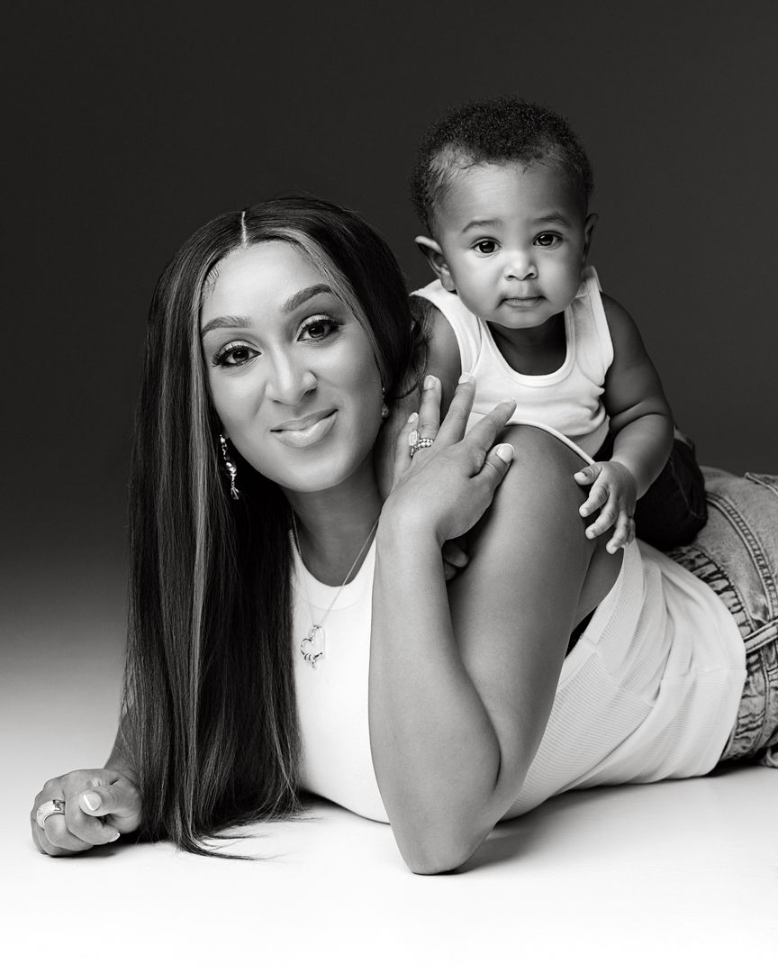 Black and white portrait of a smiling woman lying down with an adorable baby sitting on her back in a studio.