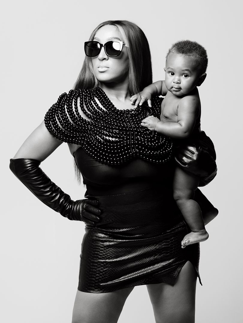Black and white photo of a stylish woman in leather outfit and sunglasses holding a baby.