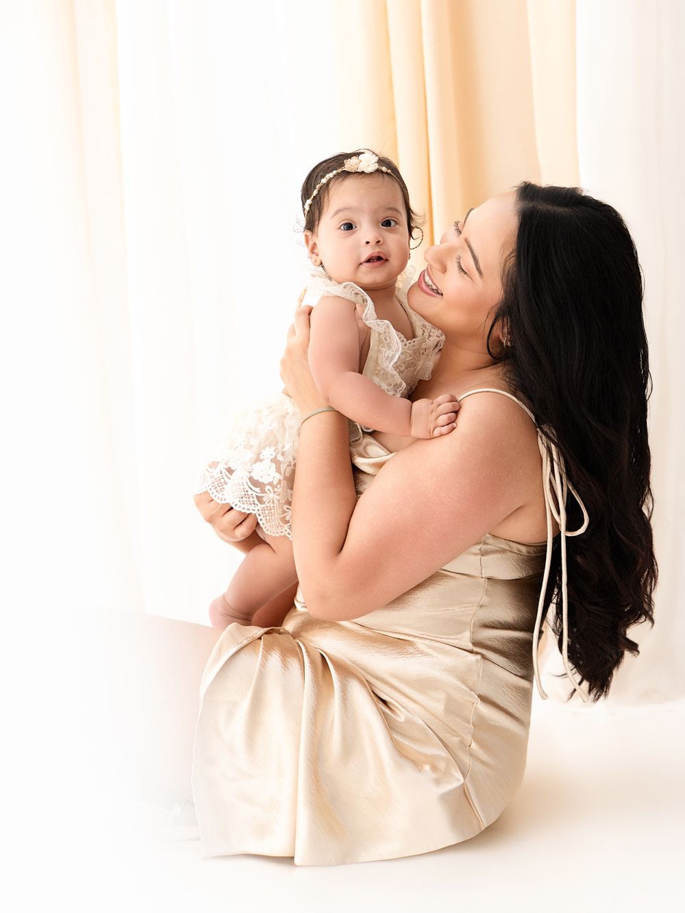 A mother in a gold satin dress sits holding her baby girl in a white lace outfit during a professional photo session.