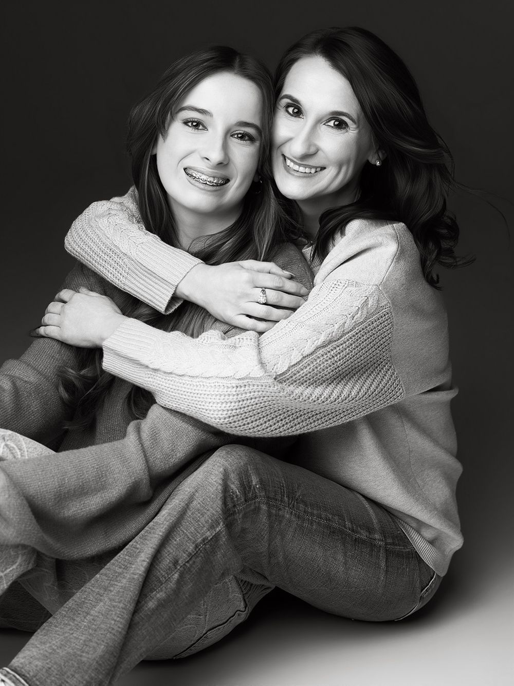 Black and white portrait of two smiling women hugging warmly, seated together in casual sweaters and jeans.