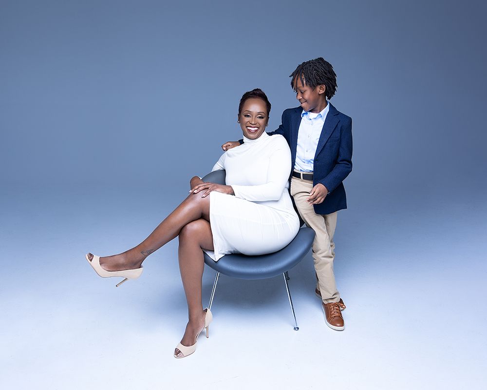 A stylish couple poses in a studio, woman in white dress seated on chair, man in blue blazer standing beside her.