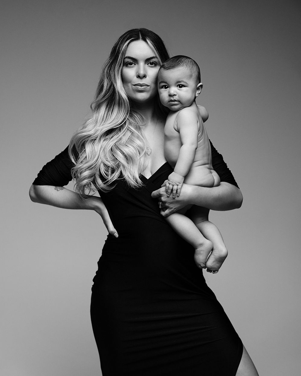 Black and white portrait of a blonde woman in a black dress elegantly holding a baby against a studio backdrop.