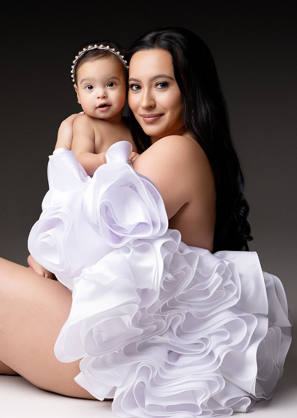 A mother in a white ruffled gown holds her baby with a pearl headband in a professional studio portrait.