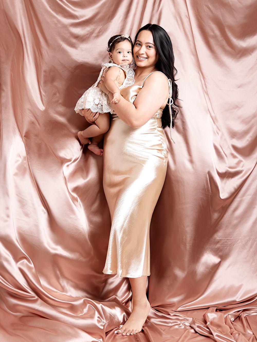 A woman in a gold satin dress holds a baby in a white outfit against a pink satin backdrop.