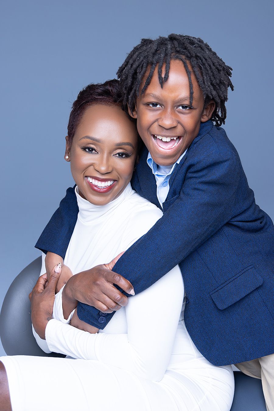 A joyful woman in white and a smiling boy in a navy blazer share a warm embrace against a gray background.