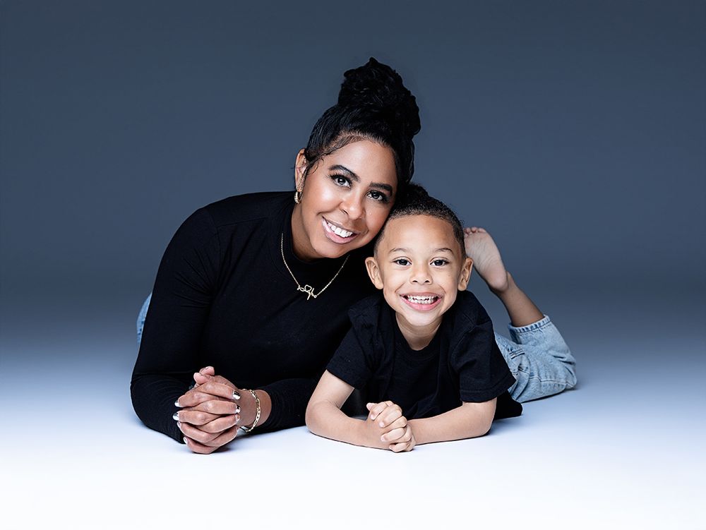 A woman and young boy smile together in matching black outfits against a studio backdrop.