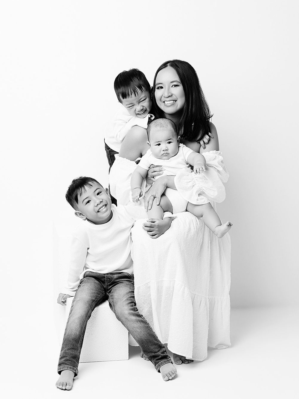 A tender black and white portrait of a family in white clothing against a plain studio backdrop.