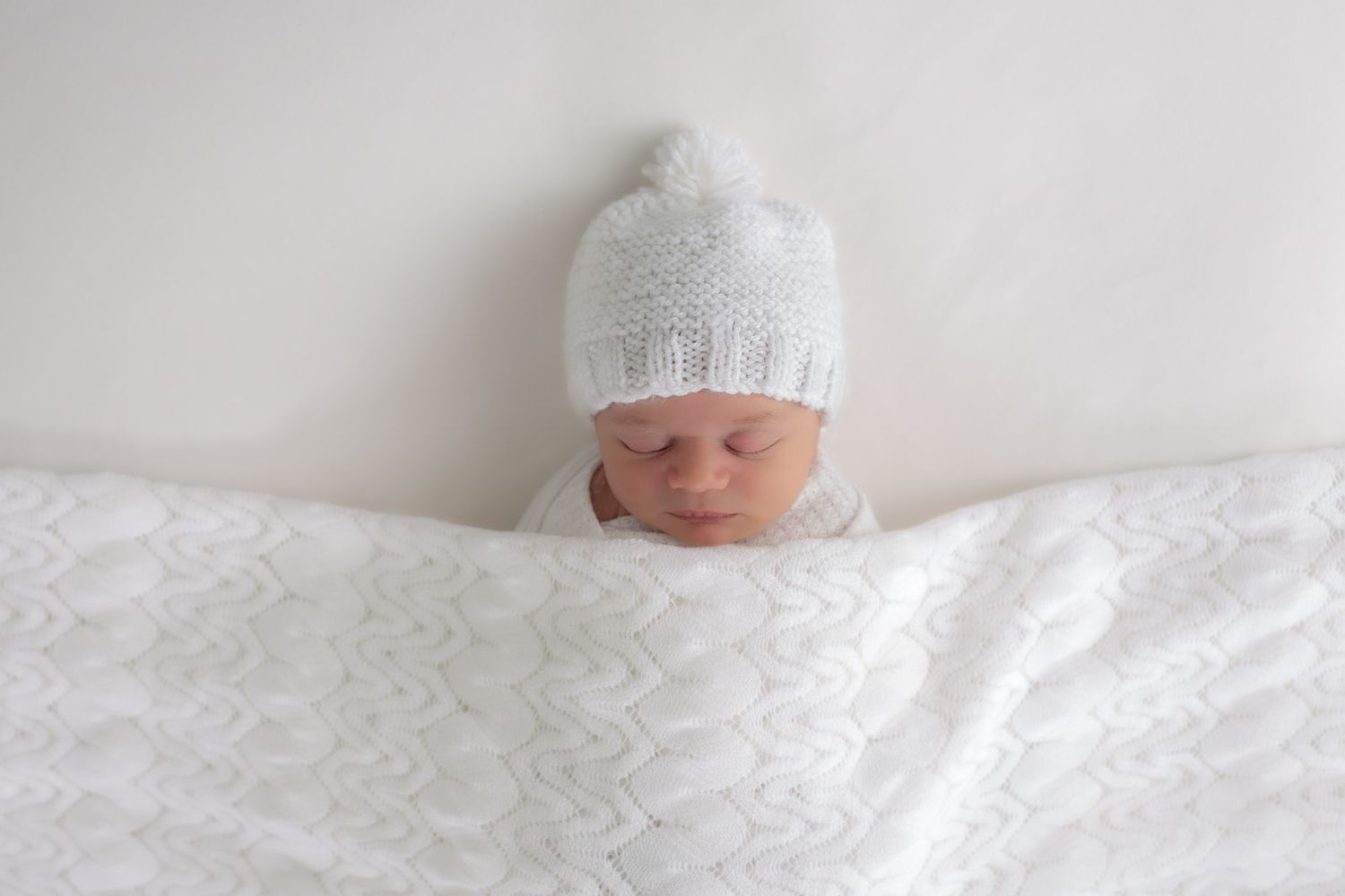 A newborn baby wearing a white knit hat rests peacefully on white bedding.