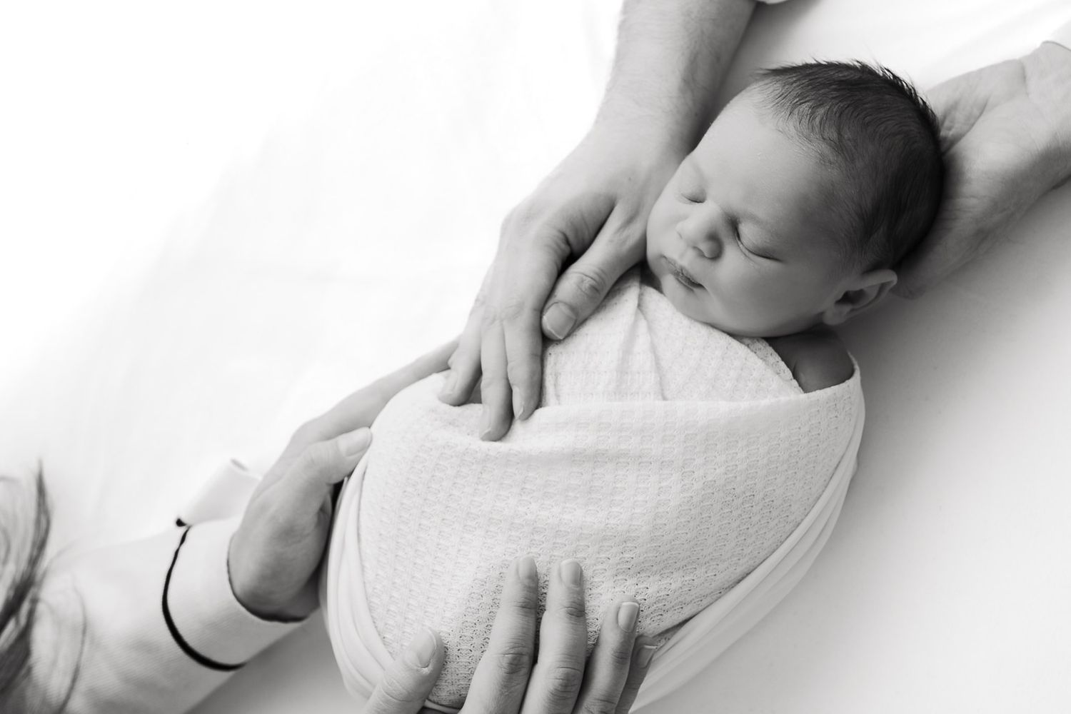 A sleeping newborn baby swaddled in white fabric is gently cradled in caring hands against a light background.